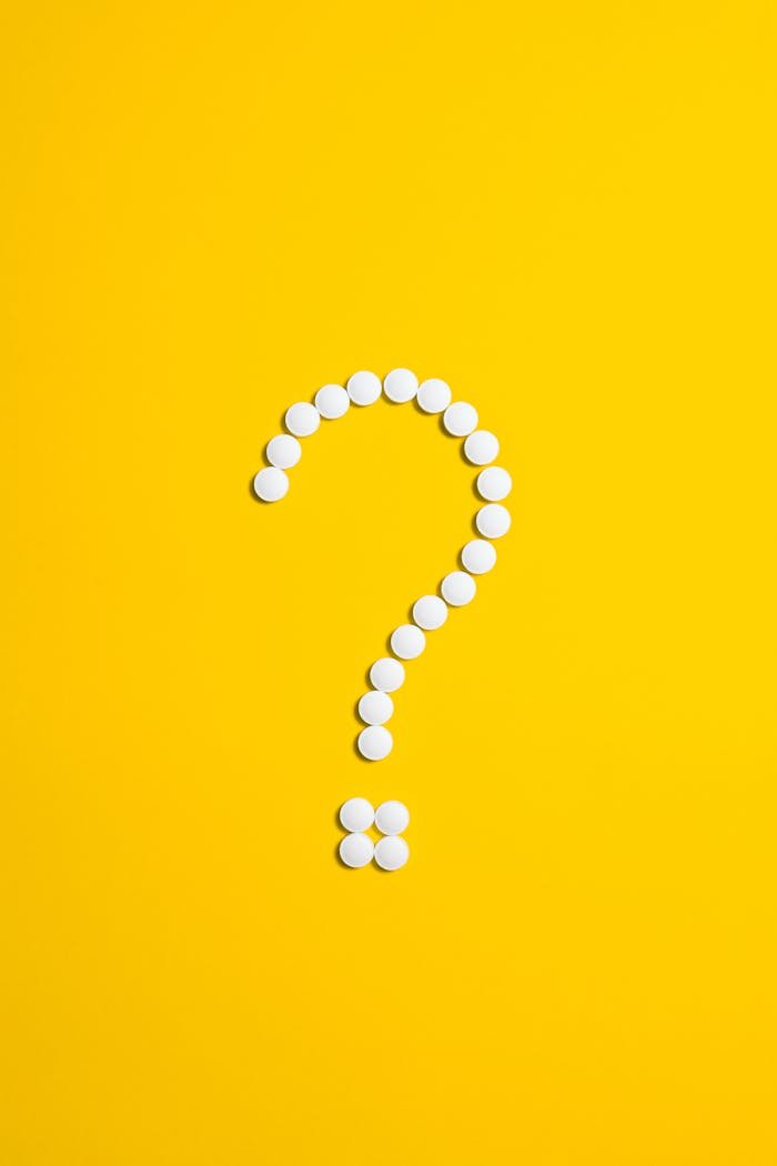 White pills arranged in a question mark shape on a bright yellow background, symbolizing medical queries.