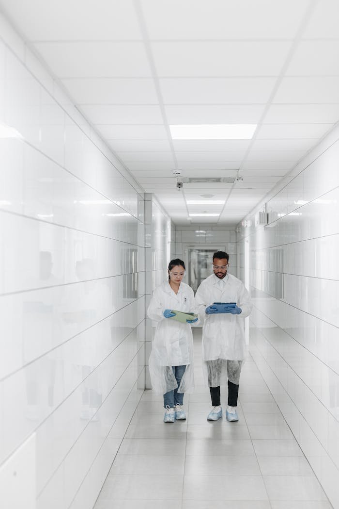 Two scientists in protective gear examine documents in a bright, sterile laboratory hallway.